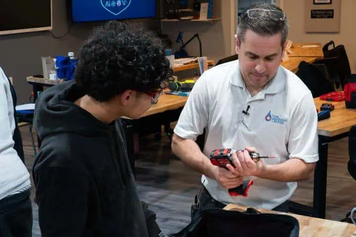 A man demonstrating a power tool to a younger person in a workshop setting