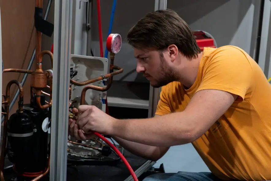 A man in a yellow shirt working on a refrigeration unit, focusing on connecting tubes and gauges.