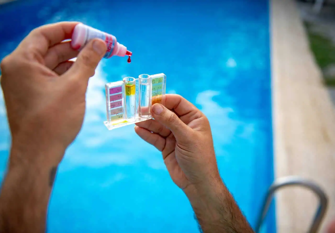 A person holding a small test kit and adding a liquid solution, with a swimming pool in the background.