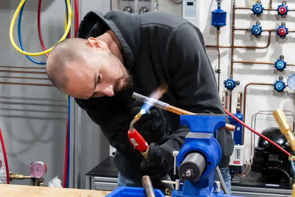A person uses a blowtorch to heat a copper pipe held in a blue bench vise in a workshop setting Image