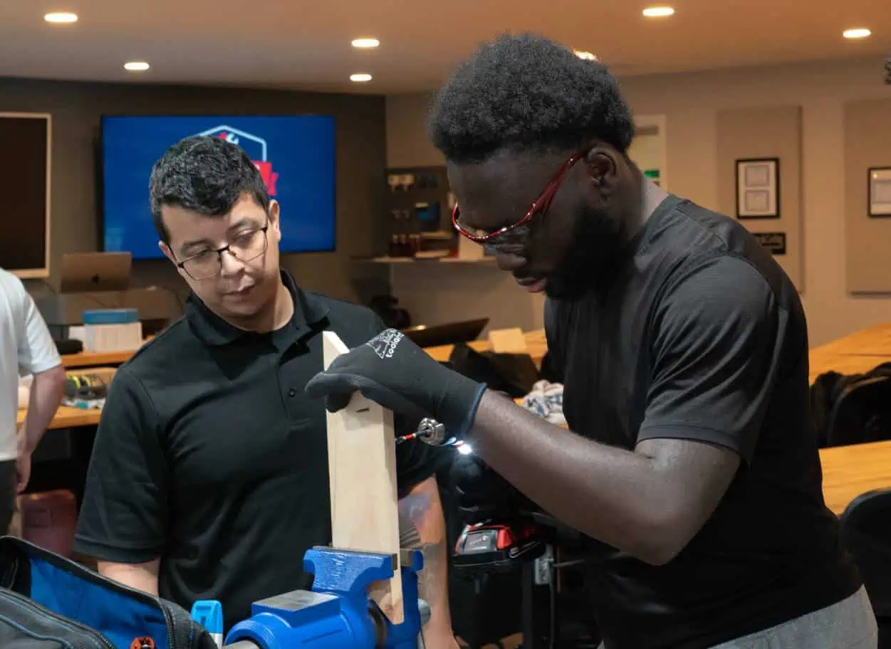 A-person-uses-a-tool-on-a-piece-of-wood-while-another-person-observes-intently-in-a-workshop-setting.
