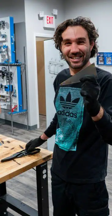 Smiling trainee holding a metal sheet at a workshop workbench with tools