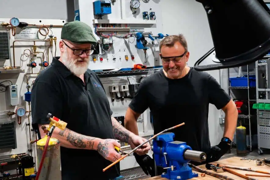 Two men cutting a metal rod in a workshop with tools around