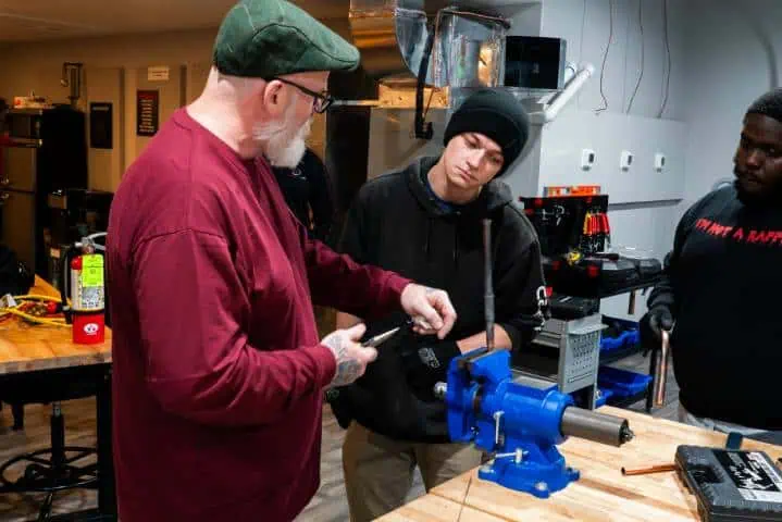 Two men working at a workshop bench, one demonstrating a tool to the other