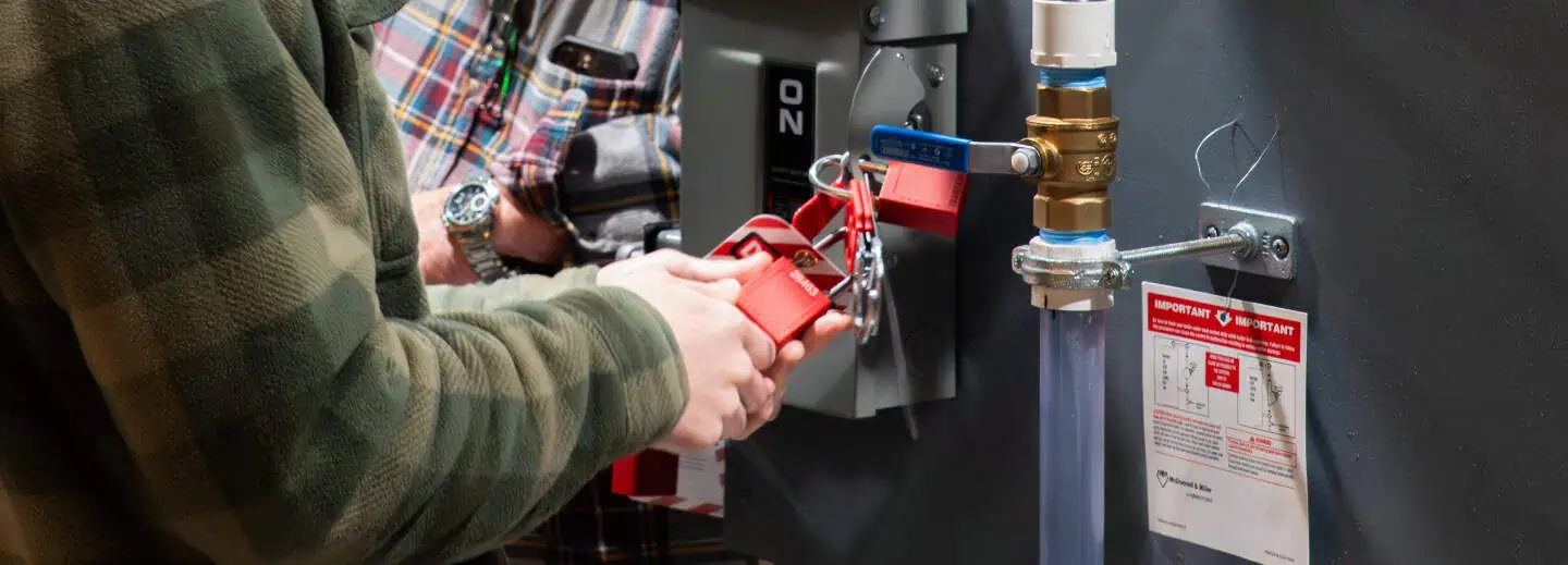 Two people are engaged in a safety procedure, applying a red lockout device to a switch, with a caution tag nearby. They are focused and attentive.