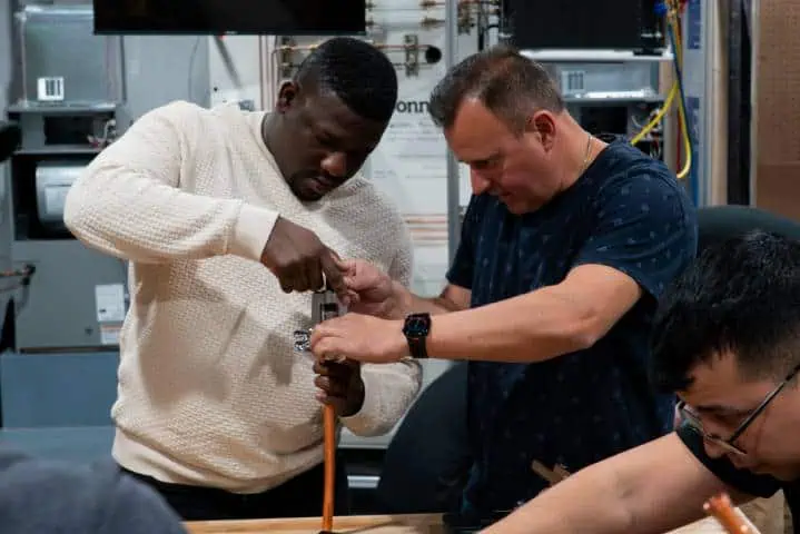 Two trainees collaborating on a hands-on pipe fitting task in a workshop.