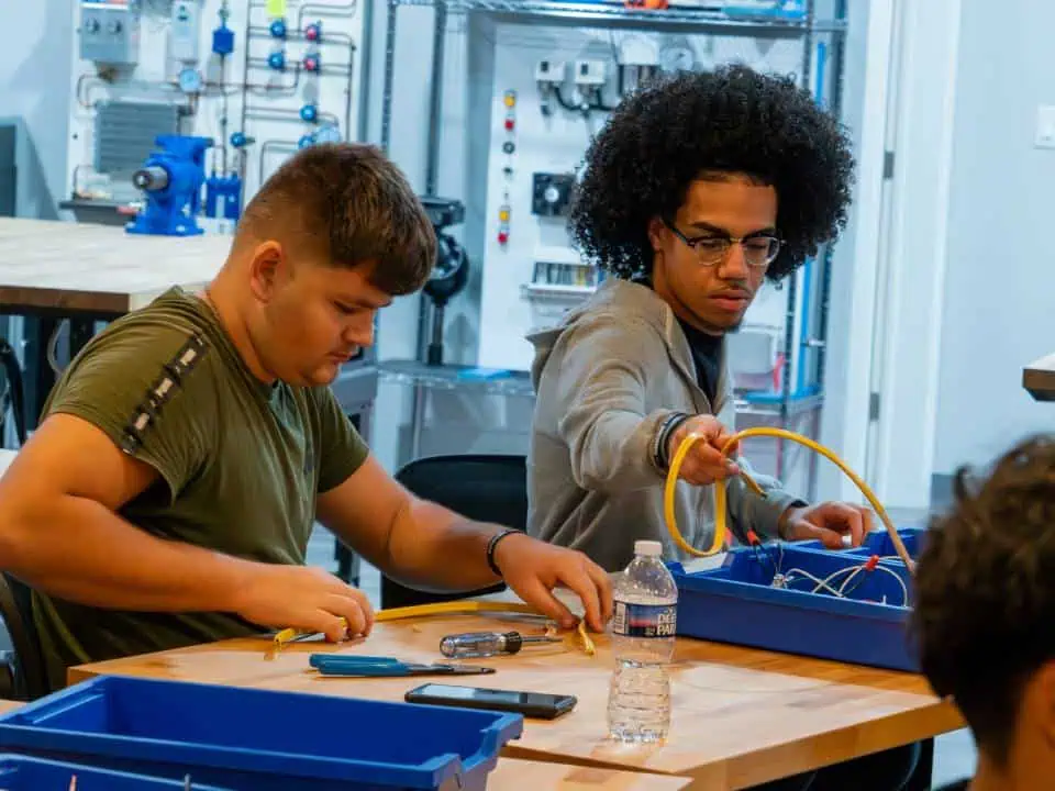 Two young men working with wires and electrical components at a workshop table